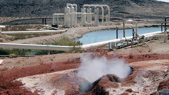 Photo of a geothermal plant with red soil and a fumarole with steam coming out of it in the foreground and hills in the background.