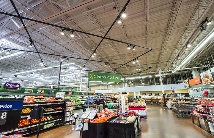 Photo of a grocery store, LED lighting hangs from the ceiling. Bins with fruit are in the foreground, a bakery on the front righthand side of the photo.