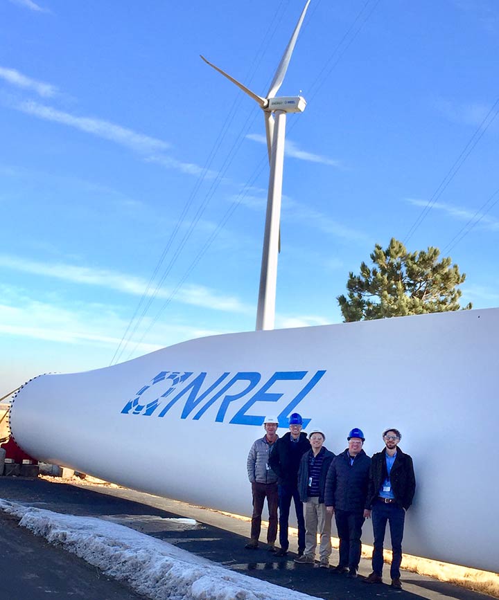 5 men wearing hard hats standing in front of a large wind turbine blade with the NREL logo.