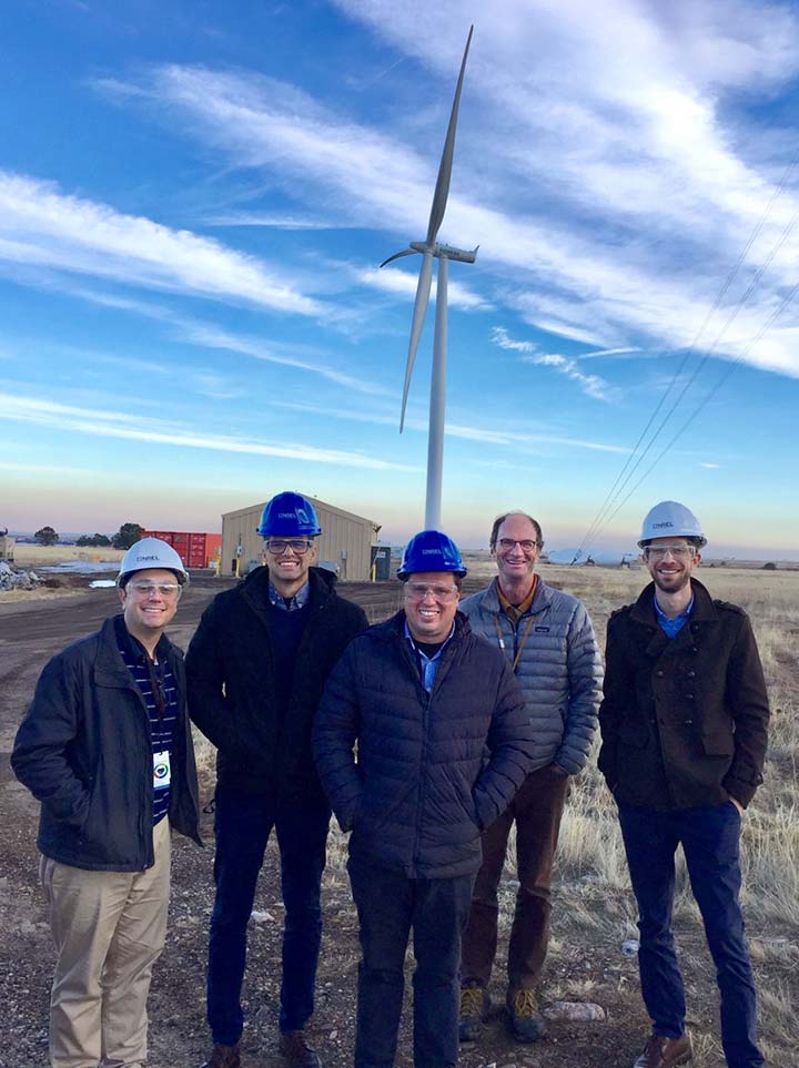 5 men standing in front of a wind turbine with their hands in their pockets (4 are wearing hard hats).