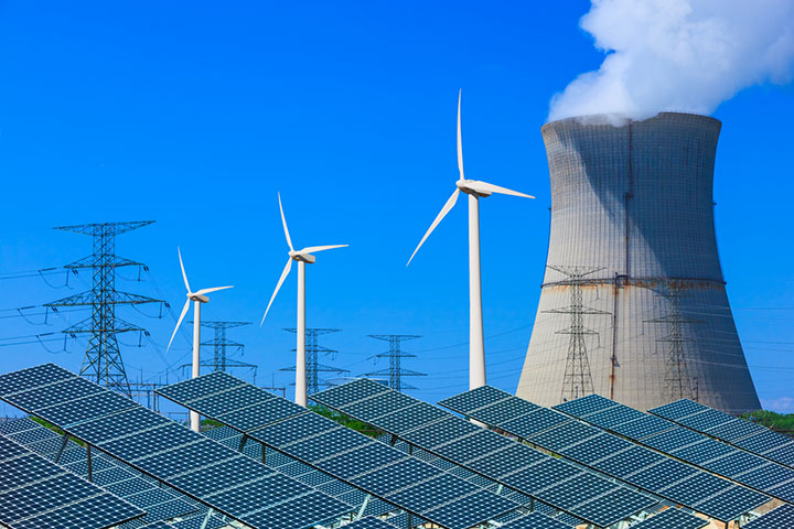 Photo of diverse energy sources, including solar panels in the foreground with a nuclear reactor, wind turbines, and power lines in the background.