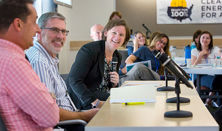 Photo of a woman holding a microphone and smiling during a meeting. She is surrounded by people smiling around a table.